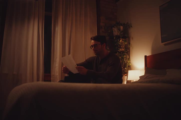 A man with glasses is sitting on a bed and reading a paper