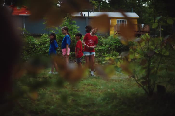 Blurred foreground of a child running away with a group of children in colorful shirts standing in a line in the background surrounded by greenery and small buildings