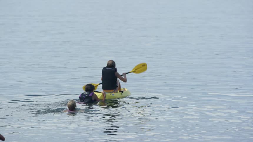 Person paddles on a kayak with two people holding onto it in the water