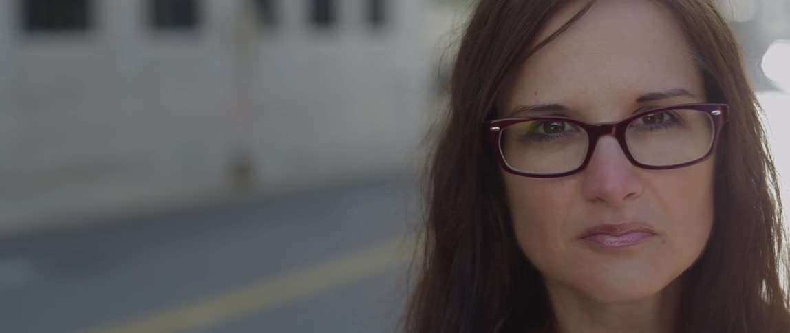 A woman in glasses standing on a road looks into the camera