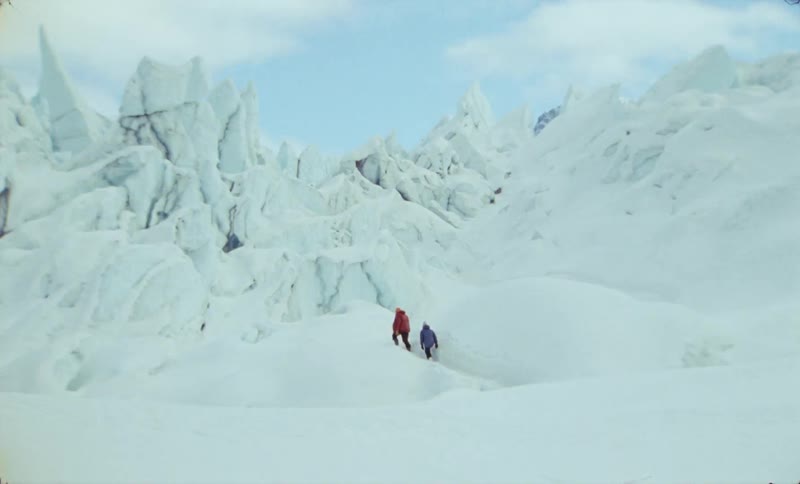 Two figures in winter clothing walk across a snowy glacier toward a background of jagged ice formations