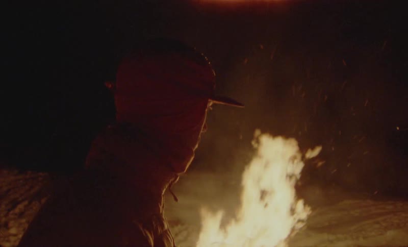 A person in warm clothes stands behind a bonfire with taller flames making lights in a dark outdoor environment