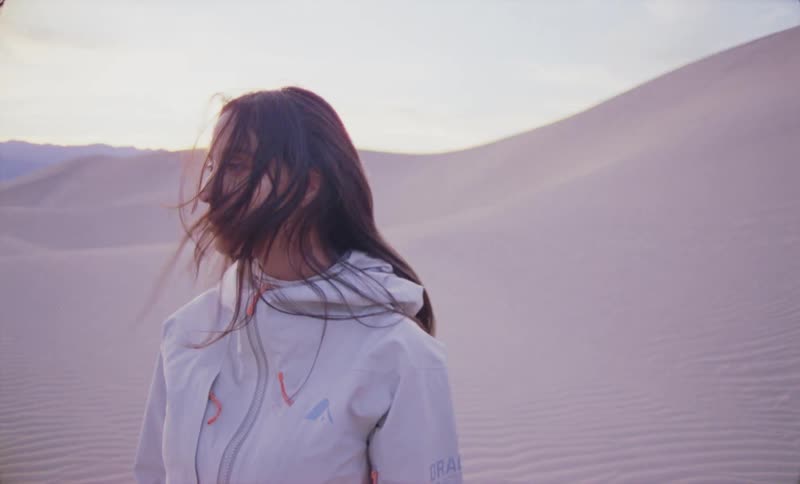 A woman standing with a white jacket and her hair is covering her face partially and she is in a desert with dunes