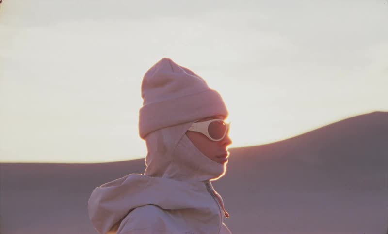 Woman standing sideways wearing a hooded coat in a mountainous landscape with the sun setting in the background