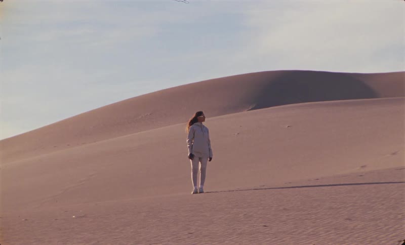 Woman dressed in light clothing stands in a vast area of sand dunes under a clear sky