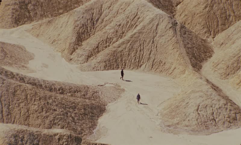 Two people walking in a dry environment with ruins and desert mountains