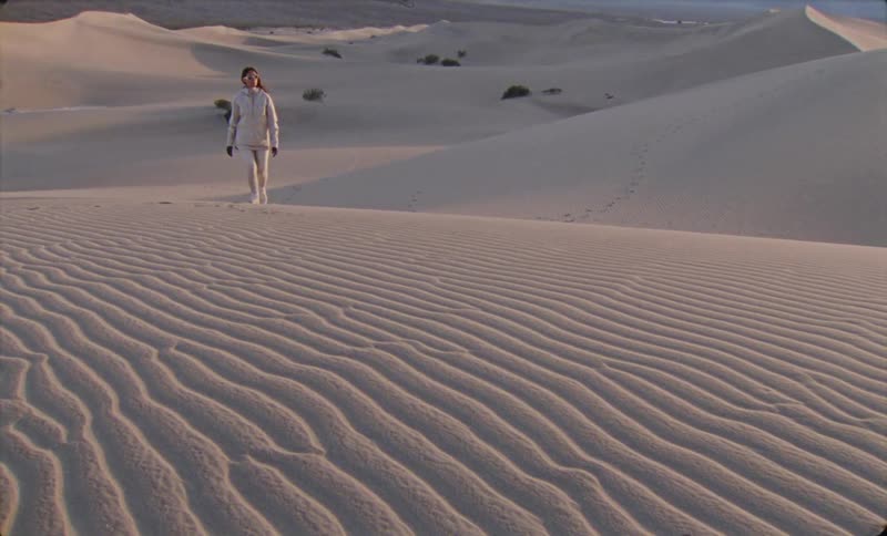 A woman in light colored clothing walks across a vast undulating sand dune landscape under a clear sky