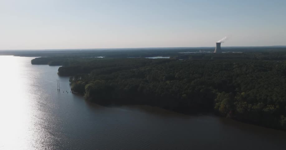 Aerial view of a river next to a forested area and a factory chimney releasing smoke in the background