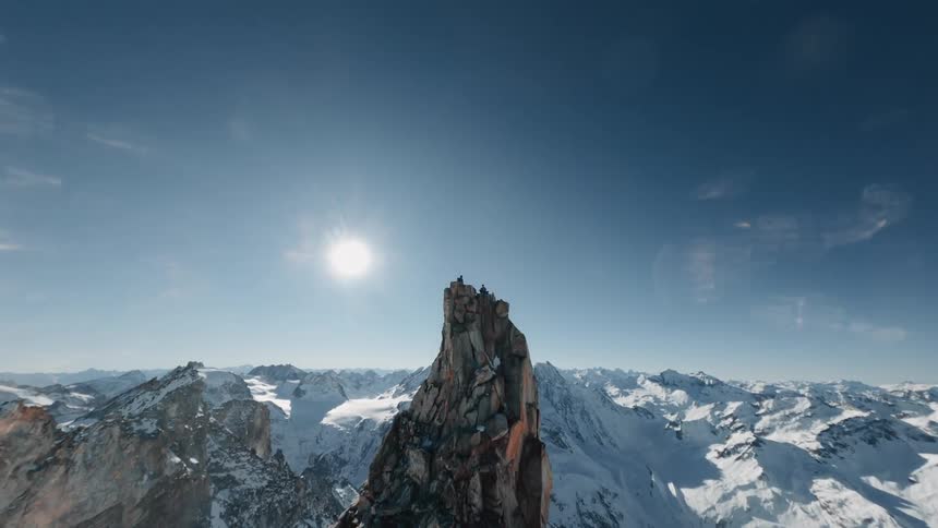 Two people on the peak of a mountain that is very high in a mountain range with snowcapped mountains filmed with a drone