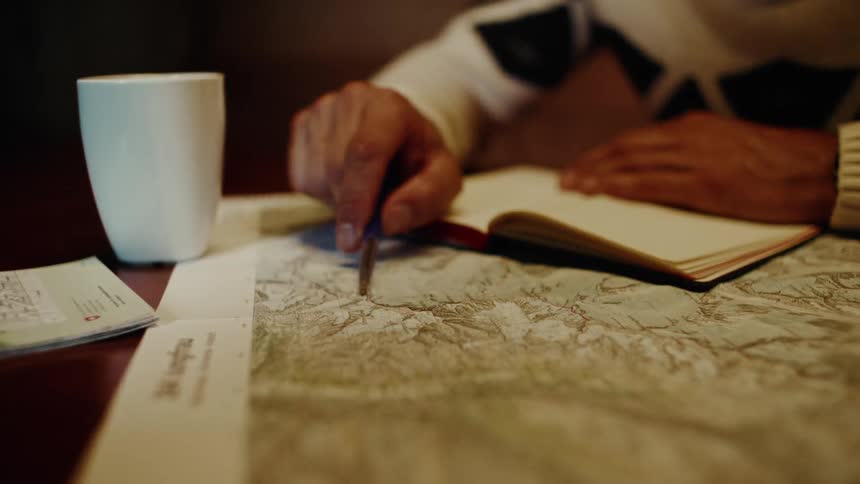 A hand uses a pencil to navigate a map on a table accompanied by a white coffee mug