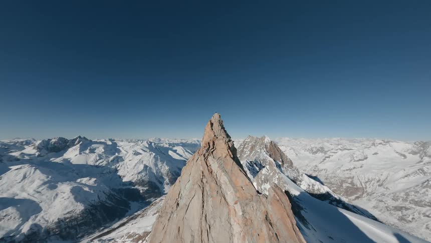 Drone shot of a person standing atop a rocky mountain peak overlooking a vast expanse of snow covered mountains under a clear blue sky