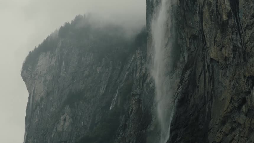 Fog hangs over a forest on top of a tall mountain with a river turning into a waterfallnearby