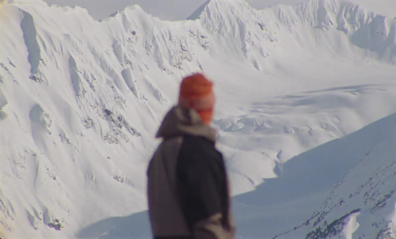 A blurry figure in a winter jacket and orange hat stands before a vast snow covered mountain range
