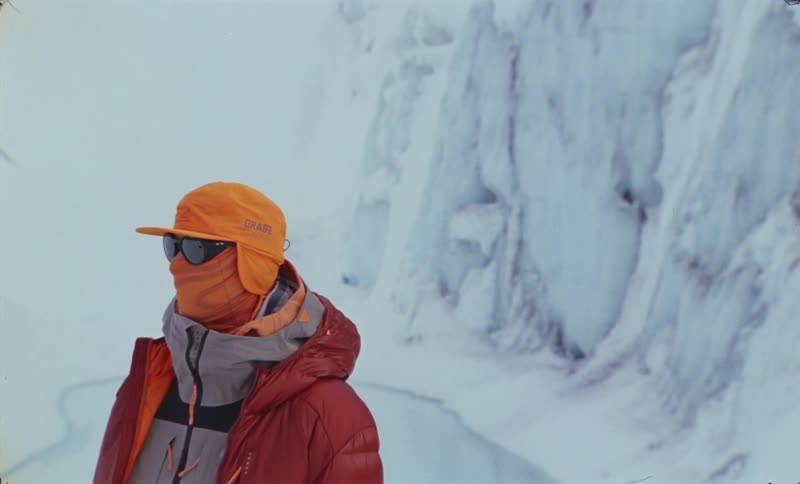 Person wearing winter clothes glasses mask and cap while standing in an environment surrounded by ice