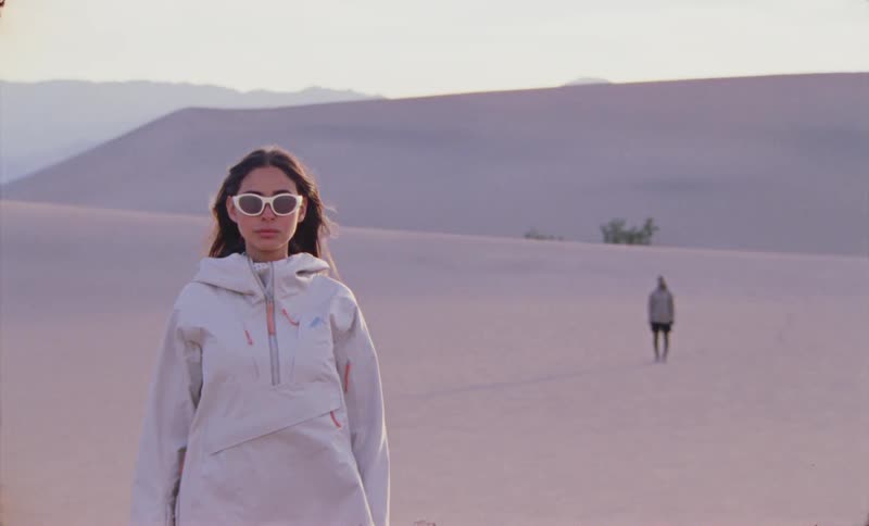 A young woman in white clothes and sunglasses is standing in a desert looking straight ahead and there is a person behind her further away