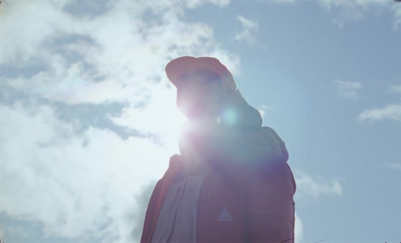 A person in heavy winter clothes and a cap is standing looking forward and back at the sky with strong sunlight
