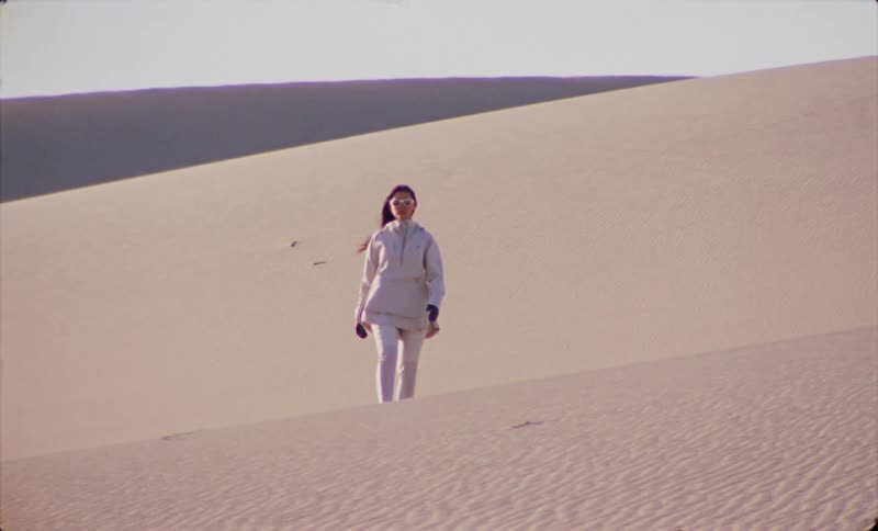 A woman wearing sunglasses walks across a desert dune