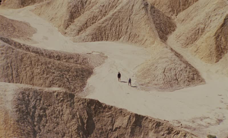 An aerial view of two people walking on a path between several sand dunes in a desert