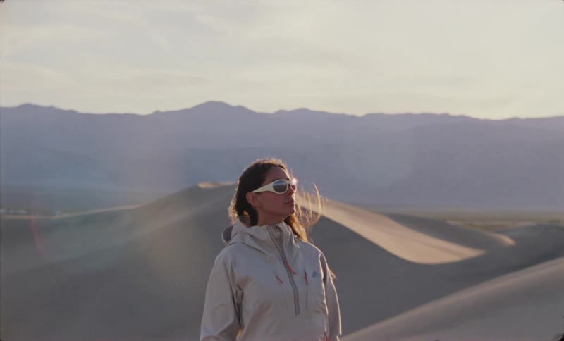 A woman stands in a windswept desert landscape wearing sunglasses and a jacket gazing into the distance