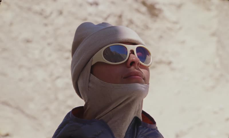A person dressed warmly with sunglasses gazes upward blending into a sandy desert backdrop