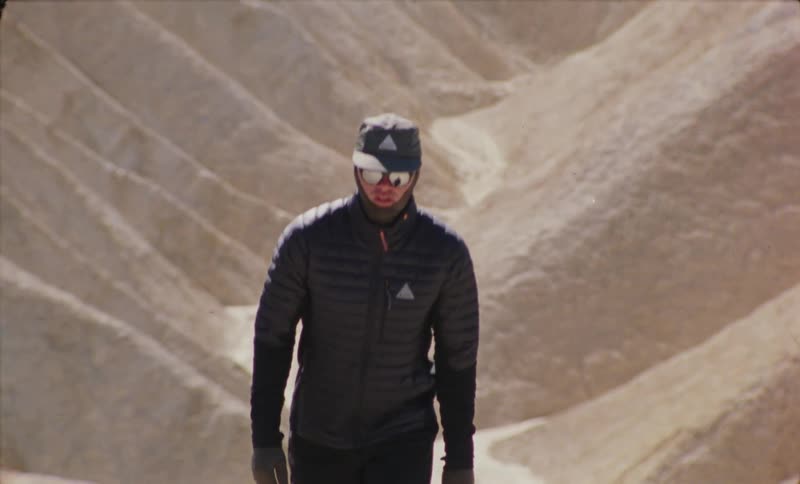 A solitary figure navigates through a rugged desert landscape wearing sunglasses and a hat