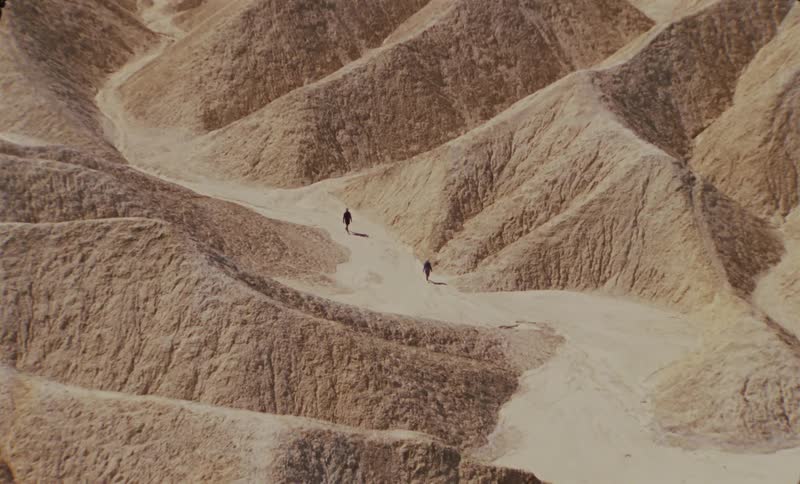Two hikers traverse a winding path through a vast arid landscape of rolling sand dunes