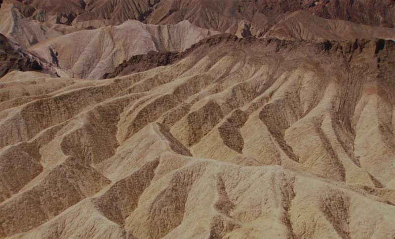 An aerial shot shows a vast expanse of tan and brown eroded desert hills and valleys