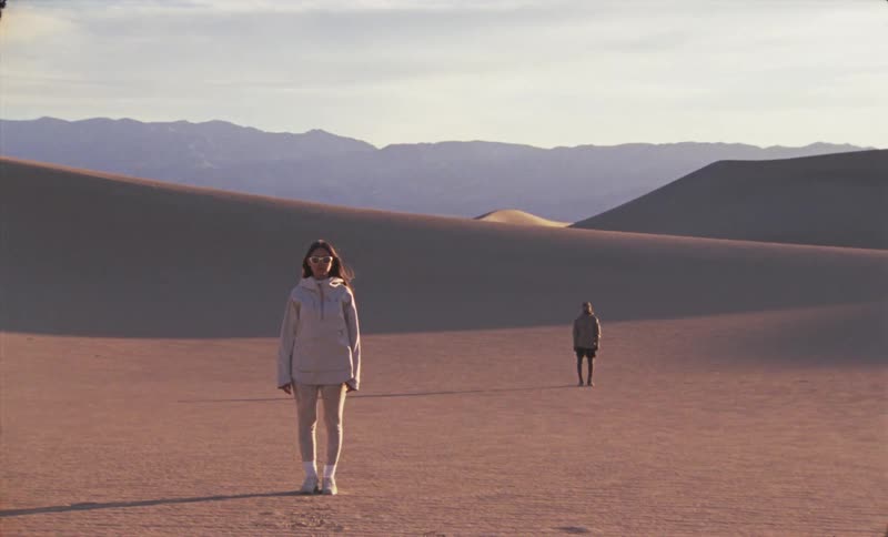 A woman in light clothing walks toward the camera in a desert landscape with a person in the distance