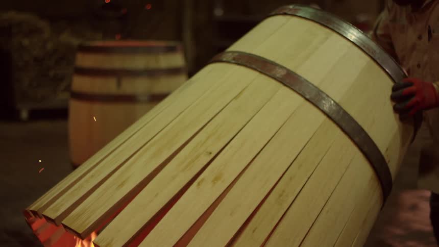 A craftsman carefully shapes a wooden barrel over a fire using a tool to heat and bend the wood