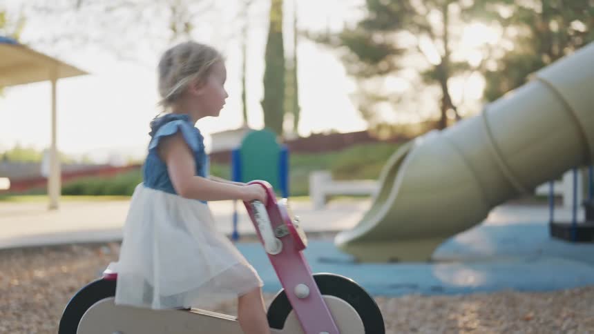 A toddler girl in a dress sits on a toy motorcycle at a playground