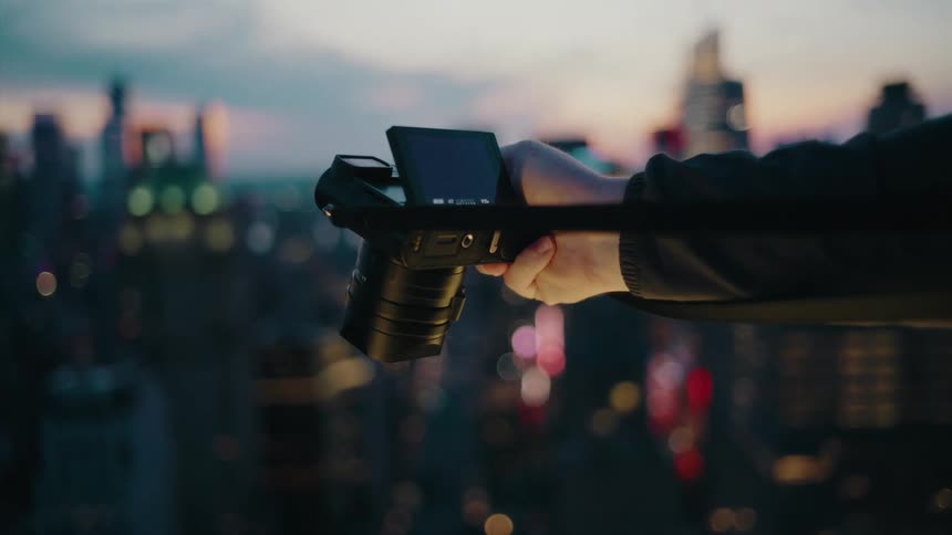 A person holds a camera with a tilted screen focusing on a blurred cityscape at dusk