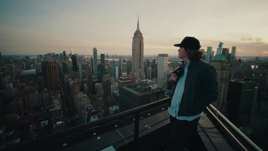A person is standing on a rooftop overlooking a cityscape with the empire state building prominently visible in the background during sunset