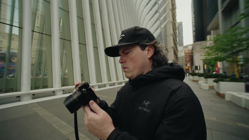 A young man in black clothes wearing a cap has a digital camera in front of his face taking pictures of something that is up and he then leaves