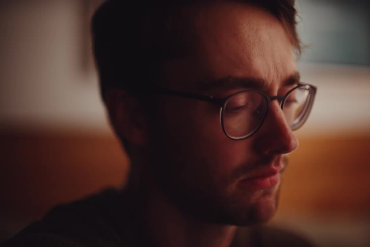 A young man with glasses appears lost in thought captured in a warm and intimate closeup