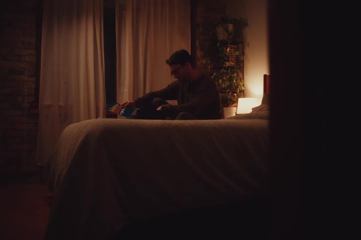 A man is sitting on a bed in a dimly lit room reading a book with a warm cozy atmosphere created by soft lighting and earthy tones