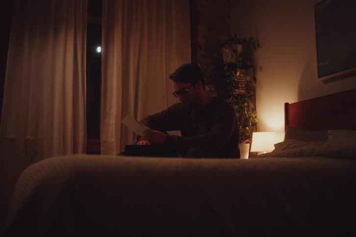 A man sitting on a bed in a softly lit room reading under the moonlight