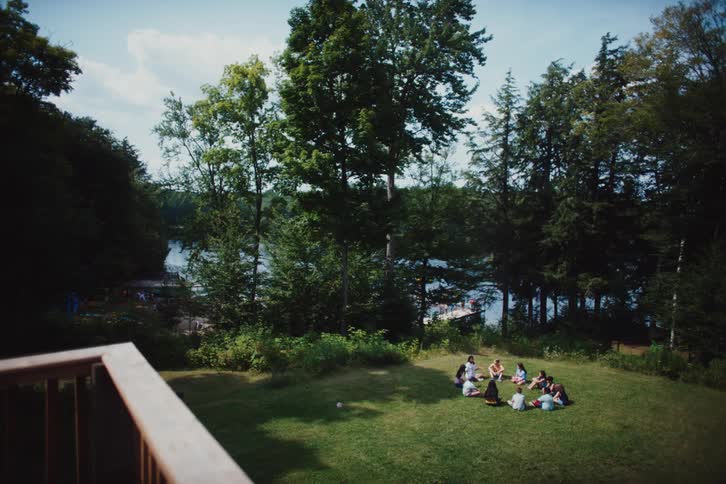 Kids gesture and talk while sitting on the lawn near a grove of trees on the riverbank on a cloudy day