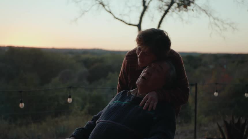 Woman kissing her husband on the cheek as he sits outside with a forest in the background