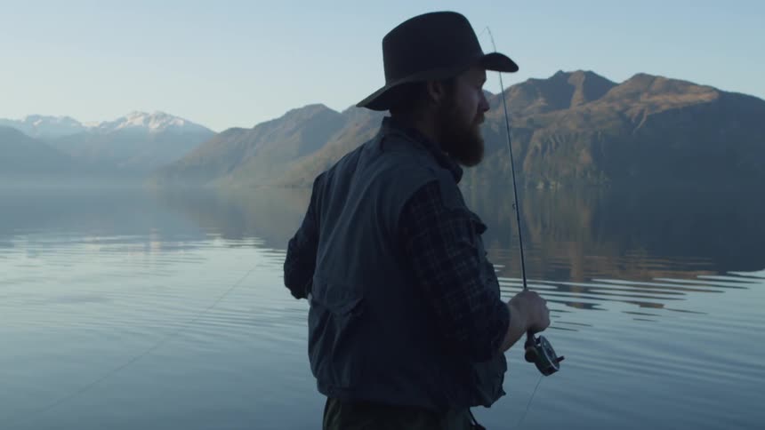 A man in fishing gear walks along the shore of a quiet lake surrounded by mountains