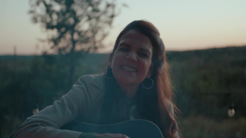 Smiling woman playing a blue acoustic guitar outdoors at dusk, surrounded by a peaceful natural setting with blurred greenery in the background.