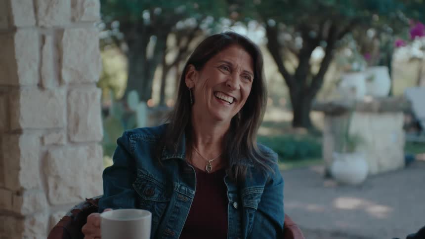 Older man and woman sit on the patio with mugs and laugh while talking