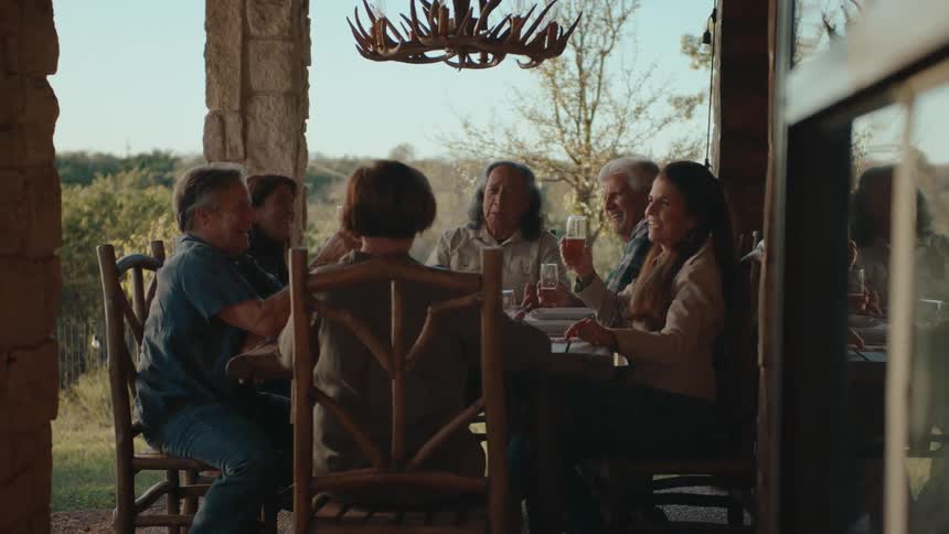 A group of people are seated around a table outdoors laughing and toasting in a covered patio area