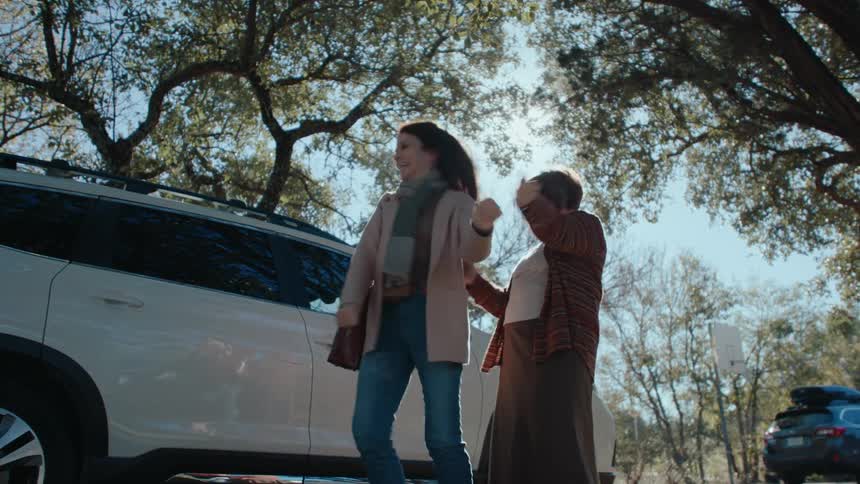 Two women one younger and one older are embracing and talking near a car with trees overhead