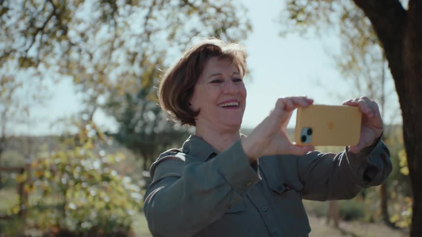 A woman takes a selfie outdoors smiling and laughing with trees in the background