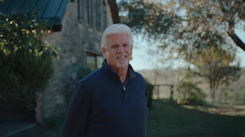 A man with gray hair and a blue shirt is in an outdoor environment standing and smiling to the front