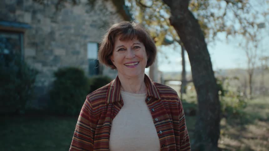 An older white woman stands in the yard of her home and smiles