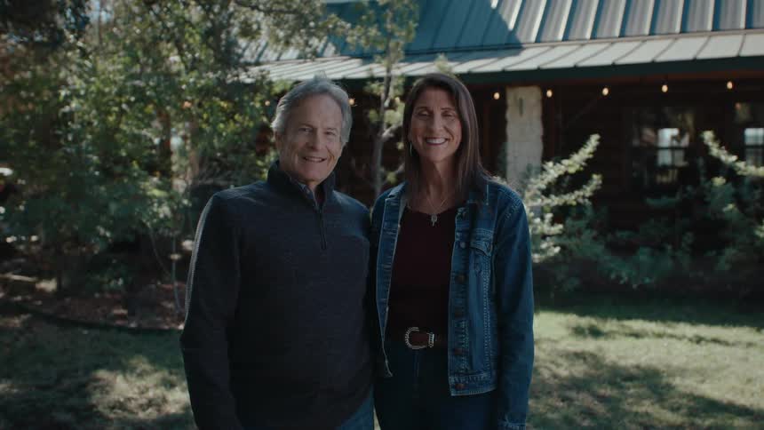 A man and a woman stand in front of a log cabin smiling and looking at the camera