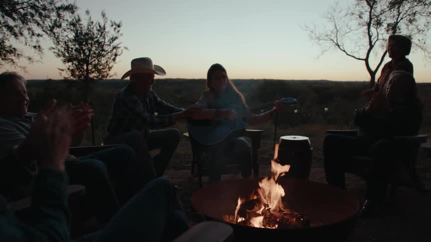 Group of people sitting around a campfire with a woman playing guitar