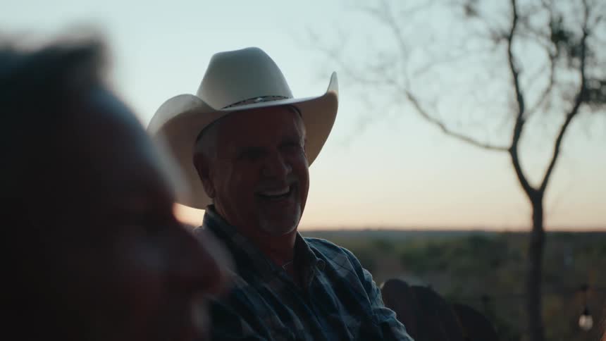A cheerful man in a cowboy hat laughs during a conversation at sunset