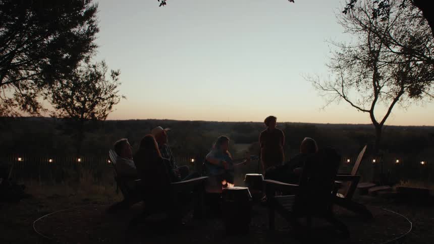 A group of people sit around a fire in a yard listening to a woman playing a guitar